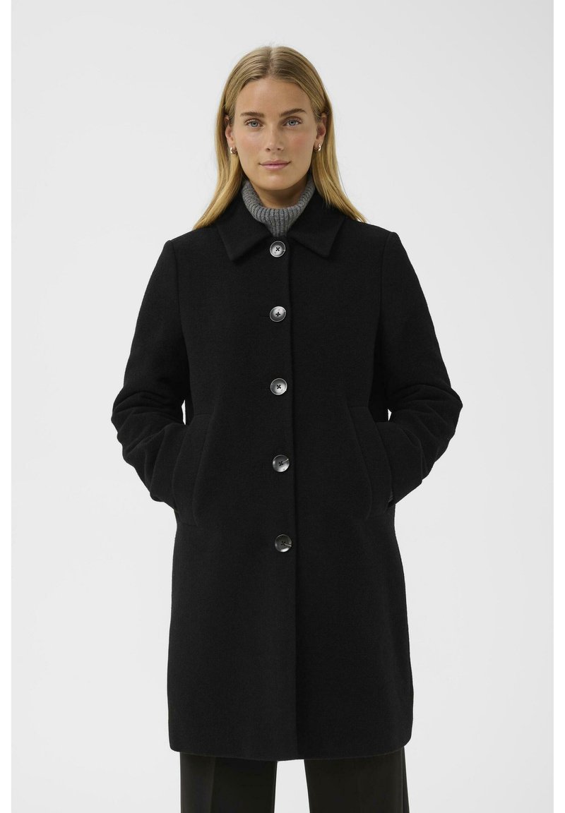 Schwarzer Wollmantel mit kragenförmigem Ausschnitt, großen Knöpfen, zwei seitlichen Taschen und taillierter Passform. Über einem grauen Rollkragenpullover getragen.