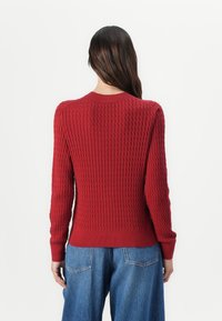 Suéter de punto acanalado rojo con mangas largas y cuello redondo. Presenta una tela texturizada y un dobladillo ajustado. Combinado con unos pantalones vaqueros de tiro alto en azul claro.
