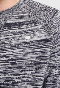 Pull en tricot présentant un motif chiné bleu marine et gris, avec un col rond et des accents côtelés sur les épaules.