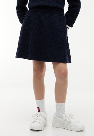 Partie inférieure du corps d'une personne portant une jupe en maille torsadée bleu marine, des chaussettes blanches avec une petite rayure rouge et bleue, et des baskets blanches.