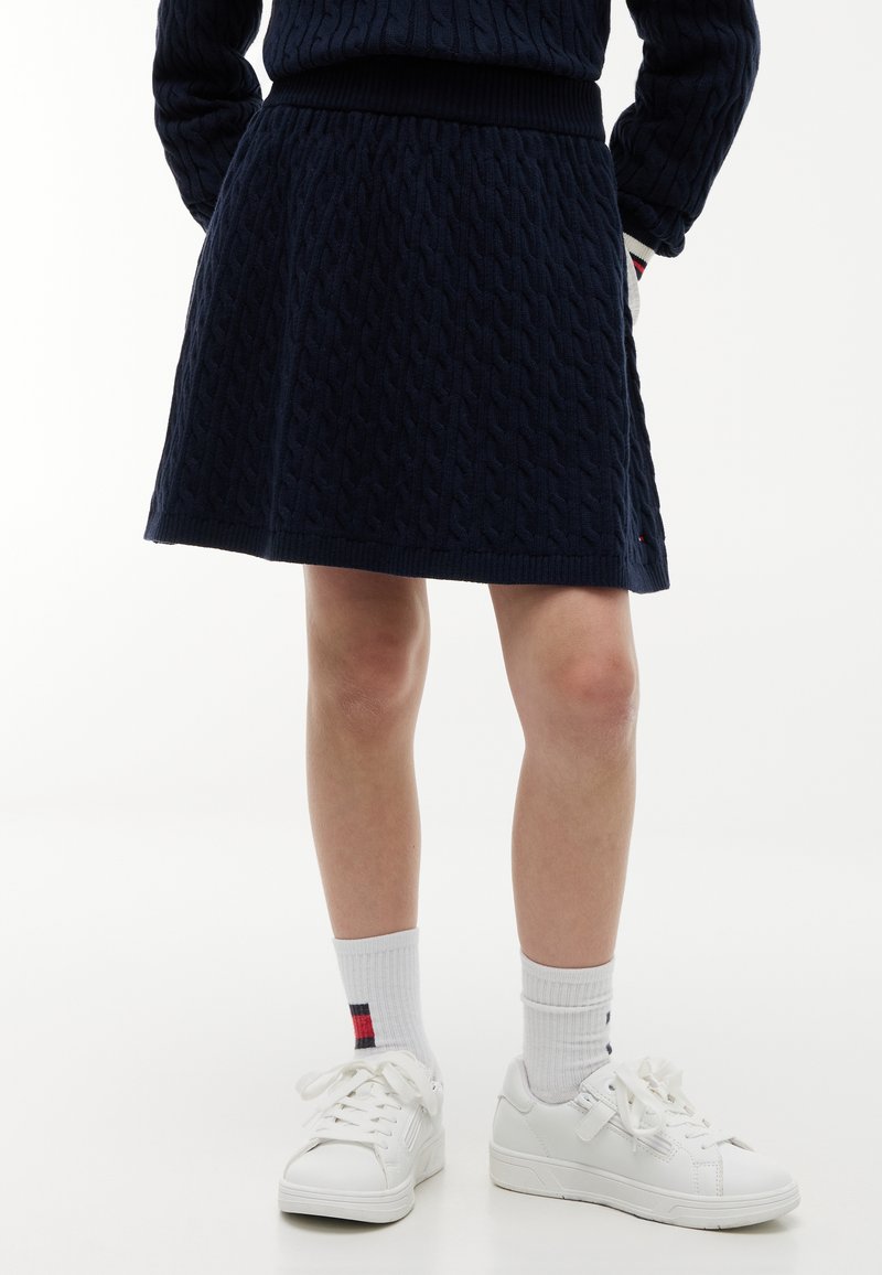 Partie inférieure du corps d'une personne portant une jupe en maille torsadée bleu marine, des chaussettes blanches avec une petite rayure rouge et bleue, et des baskets blanches.