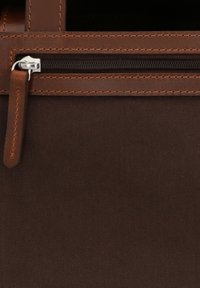 Primo piano di una sezione di borsa in tessuto marrone scuro e pelle con cerniera visibile e linguetta in pelle sul cursore della cerniera.