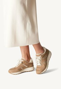 Beige suede sneakers featuring white laces, fur-lined collar, and contrasting gray stripes, with a cushioned sole and textured grip.