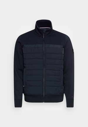 Veste bleu marine à fermeture éclair avec panneau avant matelassé, col montant, poches latérales et marquage discret sur la poitrine et la manche.