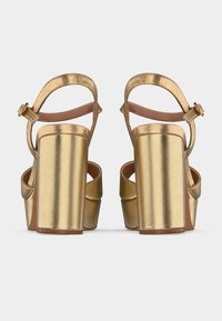 Ζευγάρι μεταλλικές χρυσές σανδάλες με χοντρά τακούνια και λουράκια στον αστράγαλο με αγκράφες, εμφανίζονται από πίσω σε ανοιχτό φόντο.