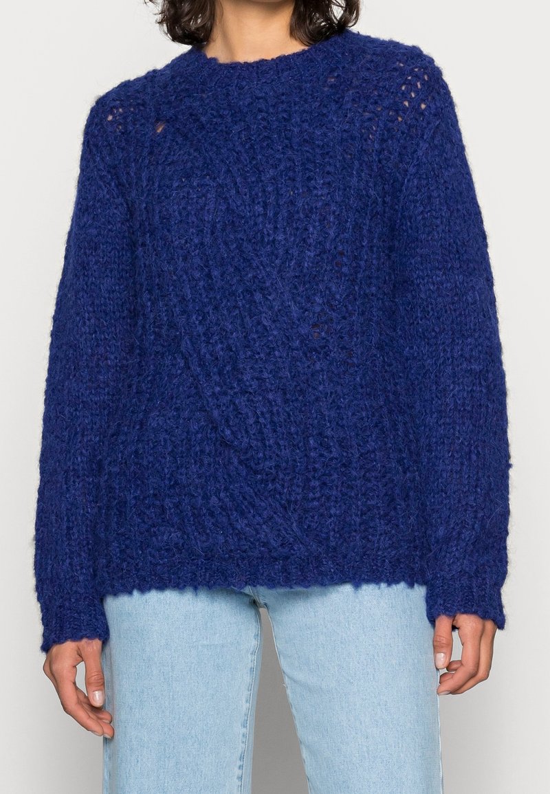 Pletený svetr v tmavě modré barvě s texturovaným vzorem, kulatým výstřihem a dlouhými rukávy. Materiál působí měkce a hrubě.