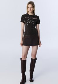 Zwarte fluwelen t-shirt met de tekst "Trust Is Fragile", gecombineerd met een bruin suède mini-rock en bruine kniehoge laarzen. Soepele texturen, aangesloten ontwerp.