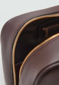 Hnedá kožená taška s hladkým povrchom, vybavená zlatým zipsom a vnútorným vreckom na zips. Okrúhly tvar a kontrastná tmavá podšívka.