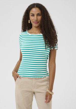 Femme aux cheveux bouclés portant un haut à manches courtes rayé turquoise et blanc et un pantalon beige, debout avec une main dans la poche.