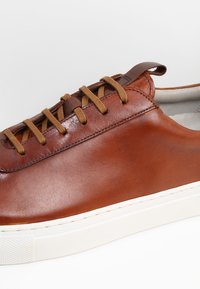 Braune Leder-Sneaker mit einer glatten Oberfläche, kontrastierenden braunen Schnürsenkeln und einer weißen Gummisohle. Verfügt über ein minimalistisches Design und gestickte Akzente.