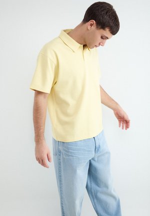 Jonge man die een bleekgeel korte mof polo-shirt en lichtblauwe spijkerbroek draagt, kijkt naar beneden met licht gestrekte armen.