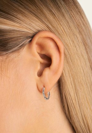 Orecchino a cerchio in argento con design intrecciato, esposto sul lobo di un orecchio, caratterizzato da una texture metallica liscia e un leggero luccichio.
