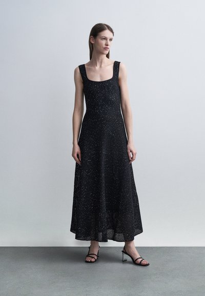 Γυναίκα που φοράει ένα μαύρο midi φόρεμα χωρίς μανίκια με λευκές βούλες και μαύρα πέδιλα με ανοιχτές μύτες, στέκεται μπροστά σε ένα απλό ανοιχτό γκρι φόντο.