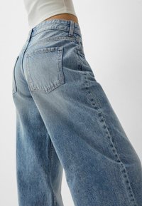 Jeans din denim ușor albastru, cu picioare largi și talie înaltă, având buzunare pe spate și o textură ușor uzată. Detalii de cusătură sunt vizibile.