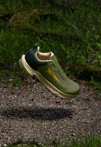 Green athletic shoe with a textured mesh upper, gold accents, cushioned sole, and a dark green heel. Suspended above a gravel path.