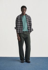 Νεαρός άνδρας που στέκεται, ντυμένος με πράσινο μπλουζάκι, ασπρόμαυρο καρό πουκάμισο, σκούρο πράσινο παντελόνι και μαύρα παπούτσια σε μπλε μοκέτα.