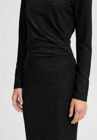 Schwarzes Kleid mit langen Ärmeln, einer figurbetonten Silhouette und einem strukturierten, glitzernden Stoff, der sich an der Taille drapiert. Sichtbare Hand, die auf dem Stoff ruht.