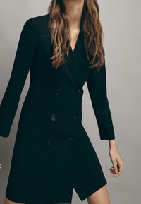 Schwarzes, doppelt ebenso geknöpftes Blazer-Kleid mit langen Ärmeln, strukturierter Form und asymmetrischem Saum. Glattes Material und Knopfakzente.