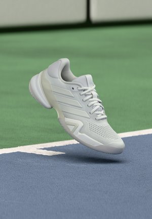 Hvit tennissko med teksturert mesh-øvre, glatte detaljer og en polstret såle, plassert over en blå banegrense på en grønn bakgrunn.