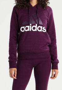 Mujer vistiendo una sudadera con capucha morada y leggings a juego, con un logo blanco y morado oscuro de "aadidas" en el frente.