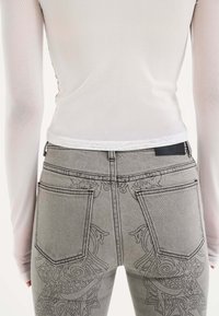 Jeans in denim grigio con intricate ricami sulle tasche posteriori, abbinati a un top a maniche lunghe bianco aderente.