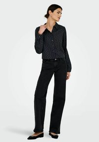 Femme portant une chemise noire à boutons avec de petits motifs rouges, un jeans slim noir et des ballerines noires avec des nœuds blancs, posant avec la main sur le col.