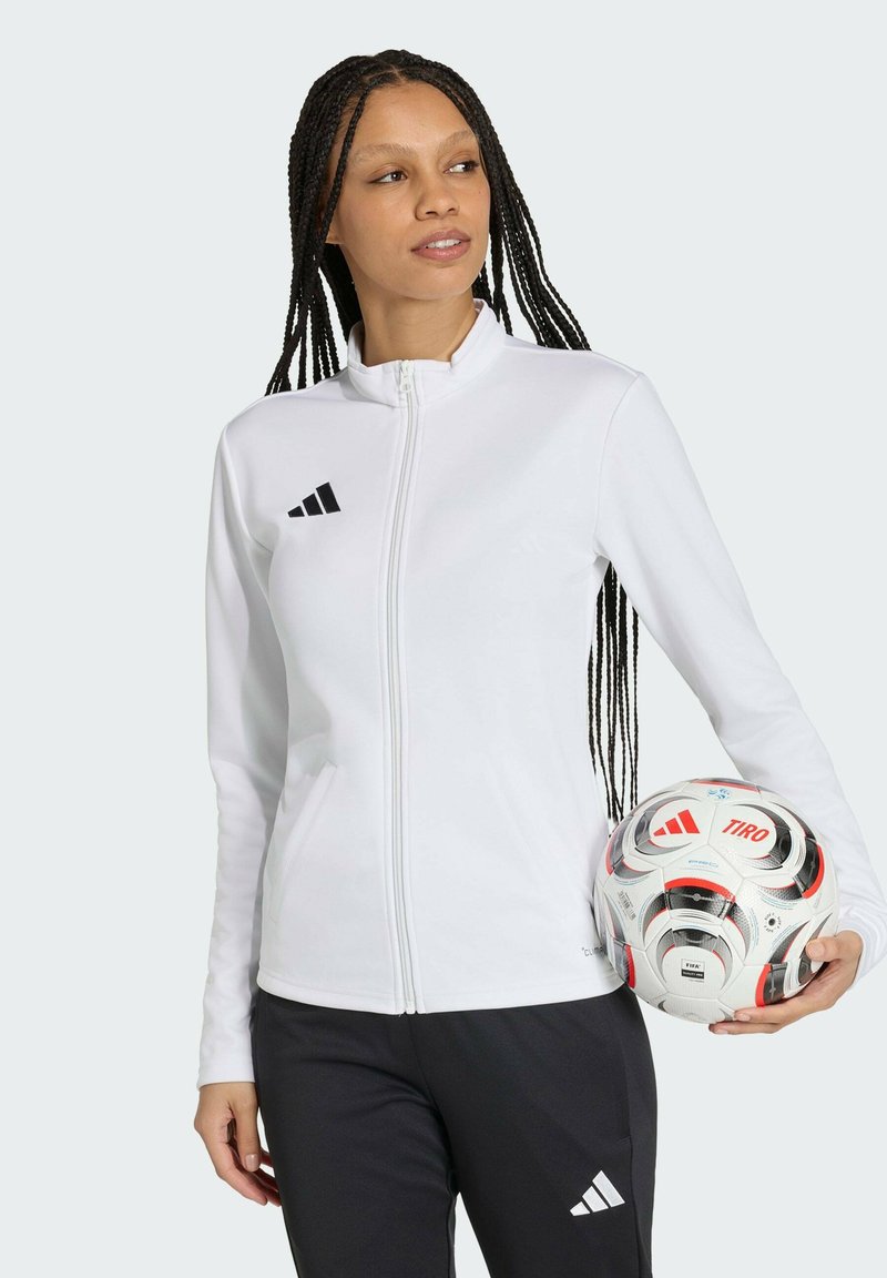 Donna con giacca bianca con zip e pantaloni neri che tiene una palla da calcio Adidas Tiro bianca, guardando di lato.