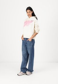 Felpa crema con design grafico rosa, vestibilità rilassata e grande tasca frontale, abbinata a jeans in denim blu larghi e sneaker in tinta chiara.
