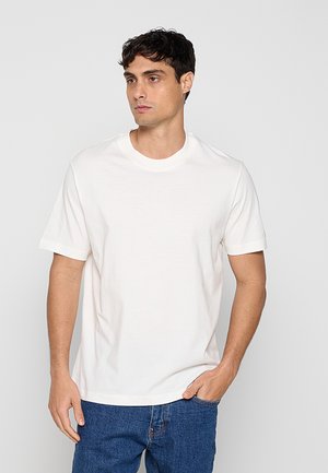 Homme aux cheveux courts et foncés portant un t-shirt blanc uni à manches courtes et un jean bleu, debout avec une main dans la poche.