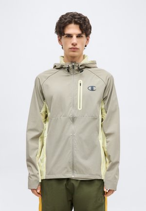 Jeune homme portant une veste à capuche zippée beige et jaune clair avec une poche zippée sur la poitrine et le logo Champion sur la poitrine.