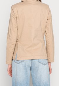 Chaqueta de algodón beige con cuello de solapa y mangas largas, que presenta aberturas laterales y un corte entallado, combinada con jeans de mezclilla azul claro.