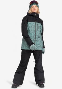 Zimná bunda s tyrkysovým vzorom a čiernymi rukávmi, doplnená o čierne snehové nohavice. Doplnky zahŕňajú rukavice a okuliare.