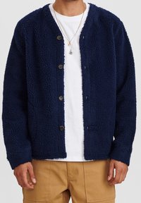 Marineblå fleece kofta med en texturerad yta, knäppning med knappar och utan krage. Har lång ärm och två framfickor.