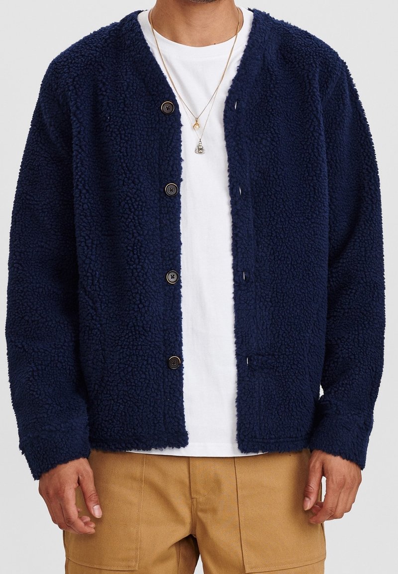 Marineblå fleece kofta med en texturerad yta, knäppning med knappar och utan krage. Har lång ärm och två framfickor.