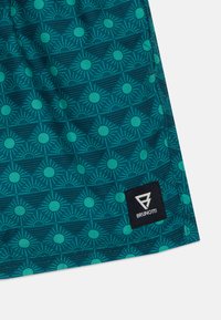 Zelena tkanina s vzorom sončnih motivov, z gladko teksturo. Vključuje črno oznako z belim logotipom v spodnjem kotu.