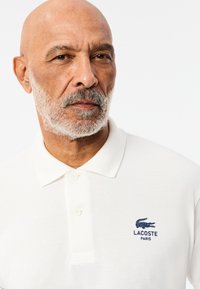 Bílá polos tričko vyrobené z texturované látky, s tří knoflíkovou légou a námořnickým logem krokodýla s nápisem "LACOSTE PARIS" pod ním.