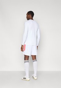 Λευκό πουκάμισο με μακριά μανίκια, λευκό σορτς με κόκκινες ρίγες, λευκές κάλτσες με μαύρες ταινίες και λευκά παπούτσια ποδοσφαίρου με κίτρινες λεπτομέρειες.