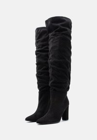 Sorte knæhøje støvler lavet af tekstureret stof, med et rynket design og subtile prikmønstre, samt en kraftig hæl for støtte.