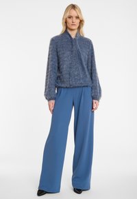 Blauer, flauschiger Pullover mit Rundhalsausschnitt und langen Ärmeln, kombiniert mit weiten blauen Hosen und schwarzen, spitzen Absätzen.