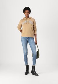 Hoodie beige avec "GANT 1949" en lettres blanches, associé à un jean skinny bleu clair et des bottines noires à talons. Accessoire sac à main vert.