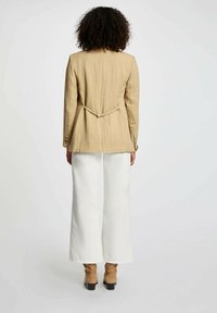 Giacca in lino beige con vita aderente, maniche lunghe e pieghe sul retro, abbinata a pantaloni bianchi a gamba larga e stivali alla caviglia marroni.