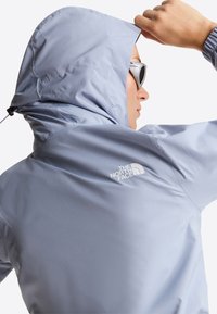 Άτομο με ανοιχτό μπλε μπουφάν με κουκούλα North Face και άσπρα γυαλιά ηλίου, που ρυθμίζει την κουκούλα, με την πλάτη προς την κάμερα.