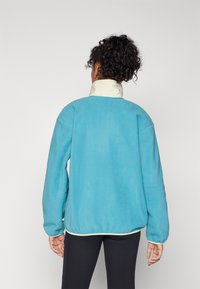 Veste en polaire bleue avec un col montant et un accent crème, dotée de manches longues et d'une coupe décontractée. Couture visible le long des bords.