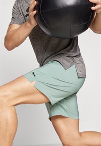 Men's light green athletic shorts with side slits, made from moisture-wicking fabric, paired with a gray short-sleeve shirt. Black logo accent.