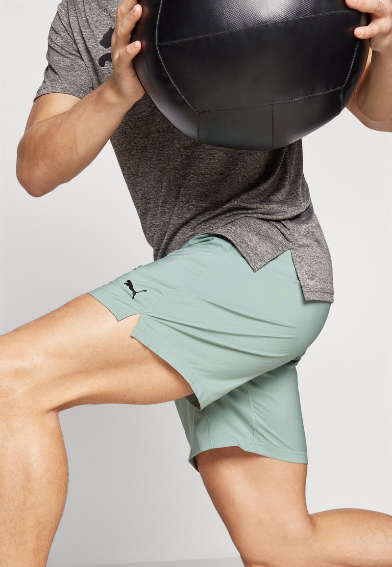 Helle grüne Sportshorts für Herren mit seitlichen Schlitzen, aus feuchtigkeitsableitendem Stoff, kombiniert mit einem grauen Kurzarmshirt. Schwarzes Logo-Detail.