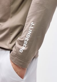 Κοντινό πλάνο μανικιού μπεζ μπουφάν με λευκή επιγραφή "J.LINDEBERG" στο μπράτσο και χέρι που ακουμπά μέσα σε τσέπη λευκού παντελονιού.