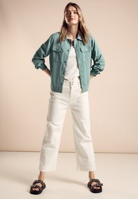 Světlá tyrkysová bunda na knoflíky se dvěma předními kapsami, bílý tričko, bílé kalhoty s vysokým pasem a širokými nohavicemi a černé sandály se sponami.