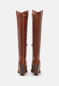Botas de cuero marrón hasta la rodilla con un acabado texturizado, tacón apilado y bordes superiores puntiagudos; cuentan con cremalleras laterales para ponerlas y quitarlas fácilmente.