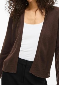 Bruine gebreide cardigan zonder knopen, open voorkant, geribbelde zoom en lange mouwen, gedragen over een witte top en zwarte broek.
