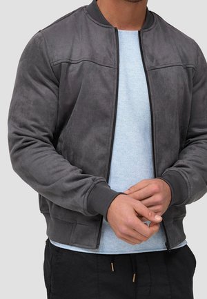 Homme portant une veste bomber en daim gris zippée sur une chemise en maille bleu clair, les mains jointes devant.